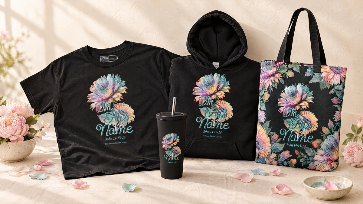 The In His Name collection together: a shirt, a hoodie, a tote, and a tumbler
