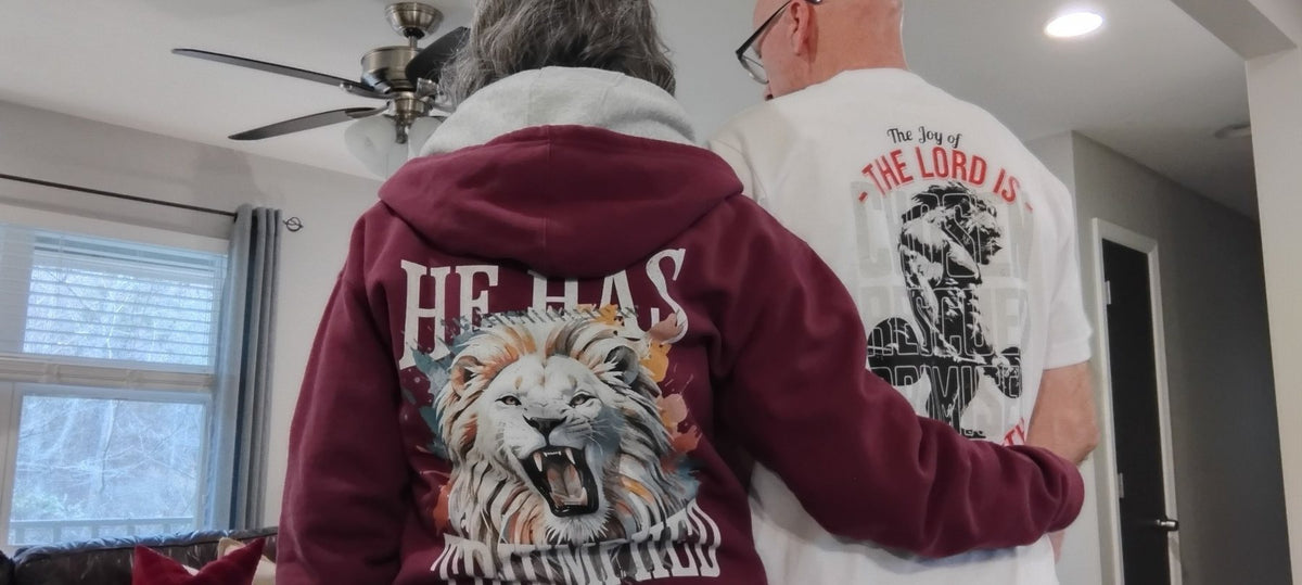 A couple in a He Has Triumphed hoodie and a Joy of the Lord tee, back prints