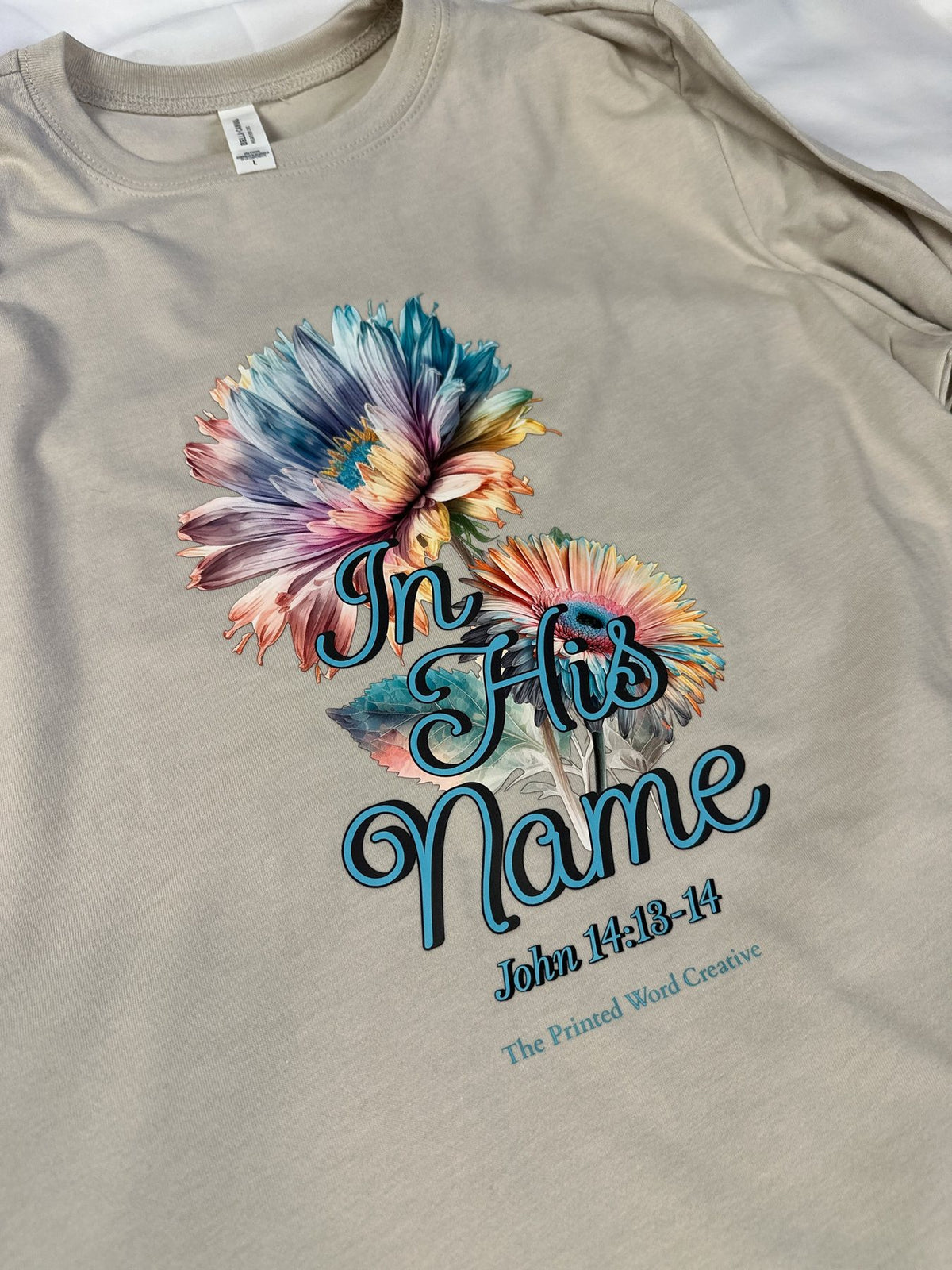 In His Name on a cream tee, laid flat